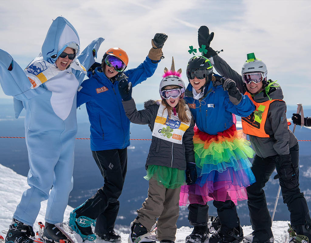 5 gals in shark costume, OAS jackets, tutus, unicorn horns and leprauchaun hats with hands in the air celebrating on mt bachelor summit