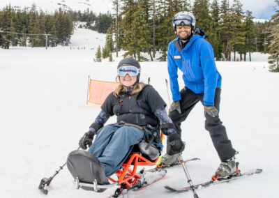 female adaptive athlete in a sit-ski smiles at the camera with helmet on, goggles down. An OAS instructor in a blue jacket stands behind smiling for the camera. The mountains and trees visible behind with a snowy landscape.