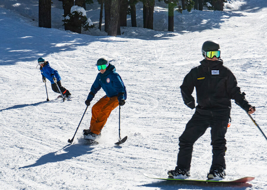 Three snowboarders are moving downslope, utilizing outriggers to provide adaptive assistance during their activity.