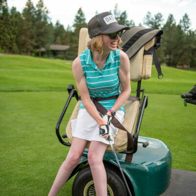 adaptive athlete in a teal polo, tan skirt, and OAS hat smiles while putting a golf ball with assistance from the adaptive golf cart.