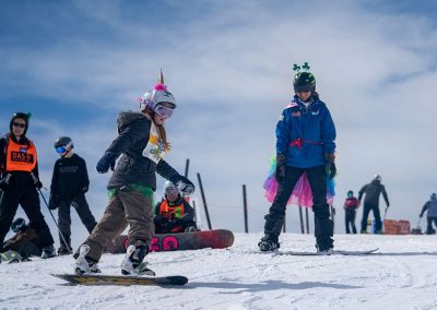 girl with unicorn horn on helmet snowboarding with instructor in costume coaching from uphill