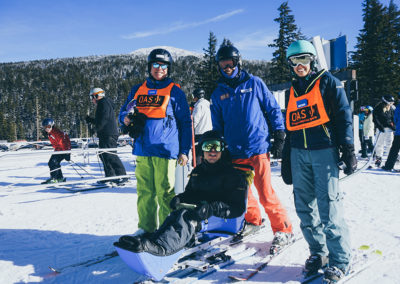 group including OAS sit skier, instructor and volunteers at base of ski lift with tumalo mountain in the background