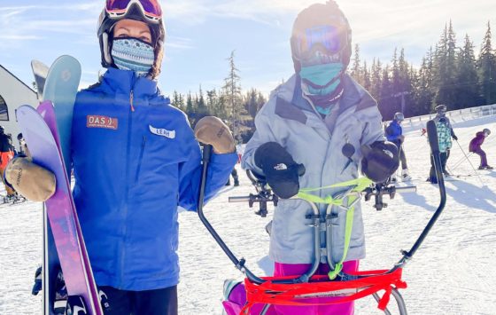 two skiers, one standing behind an adaptive ski frame