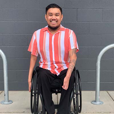 JD wearing a striped short sleeve collared shirt while sitting in his wheelchair