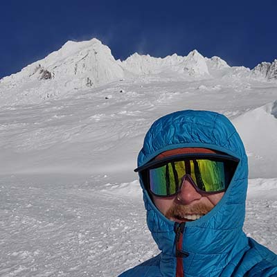 Jacob with sunglasses and a hooded jacket in front of a snowy mountain peak