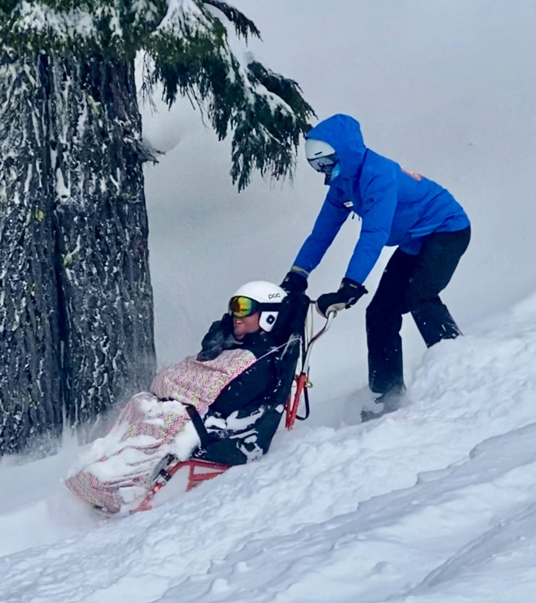 OAS instructor in a blue jacket bucketing Cory down a steep snowy terrain. Cory's smile is visible within all his winter layers and blanket from his sit ski.