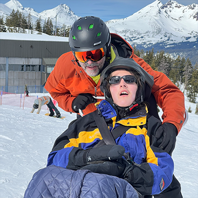 Jeff in a ski helmet and goggles standing behind his son Bryan, also wearing a ski helmet, sitting in a sit ski.