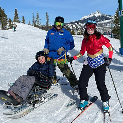 Shanna wearing a wonder woman costume standing on skis with her son Wynn in a bi-ski and OAS instructor Mike standing behind the sit ski.