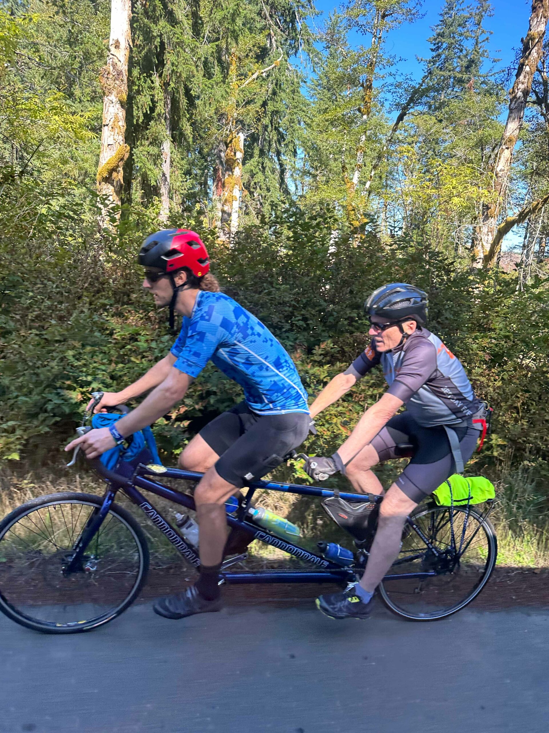 On a tandem bike there is an OAS staff, wearing a red helmet, at the front of the tandem with a blind adaptive athlete in the rear seat wearing a gray helmet.