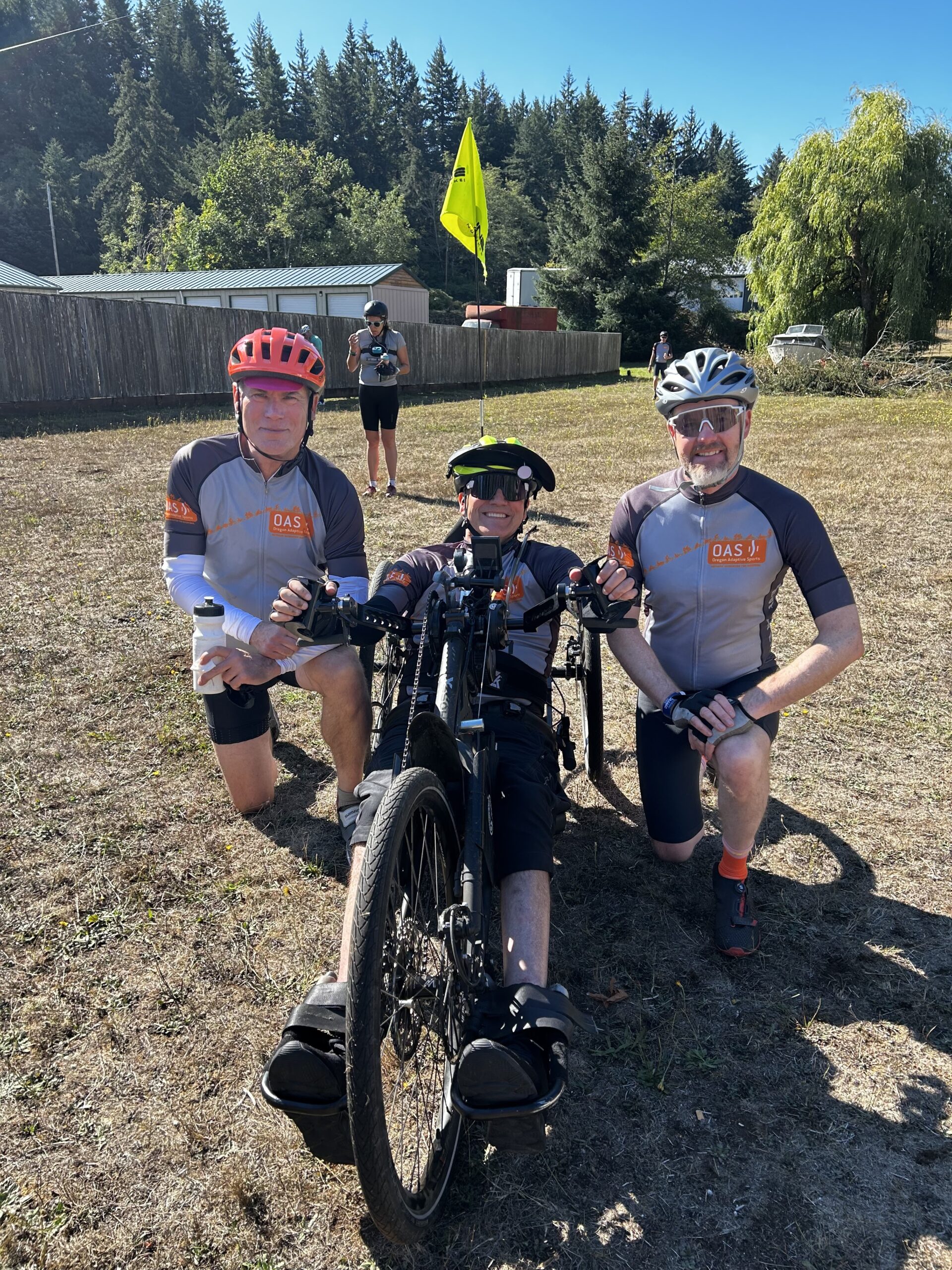 Two OAS volunteers in their OAS branded bike kits, standing beside an adaptive athlete in their hand-pedal bike