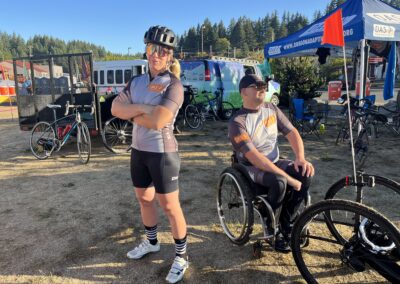 Start of the day, OAS bus and booth are in the background with two members of the OAS cycle team in front doing a power pose for the camera with each looking off to opposite sides and backs facing each other. One is an OAS Staff and another is an adaptive athlete.