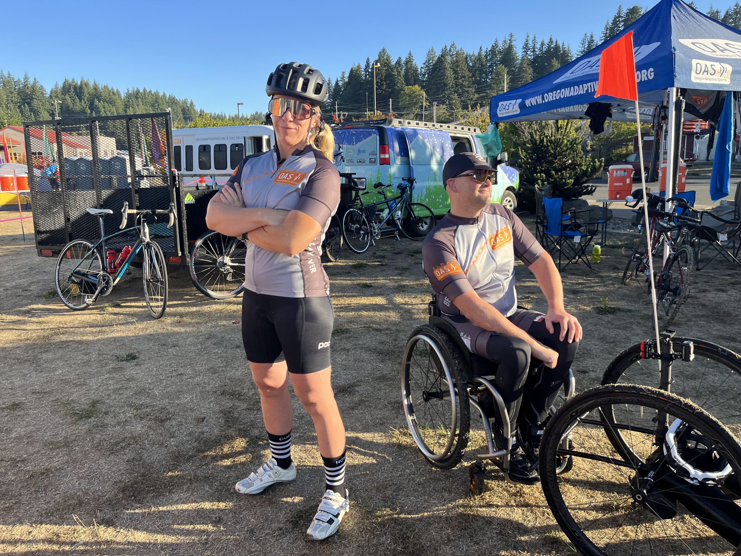Start of the day, OAS bus and booth are in the background with two members of the OAS cycle team in front doing a power pose for the camera with each looking off to opposite sides and backs facing each other. One is an OAS Staff and another is an adaptive athlete.