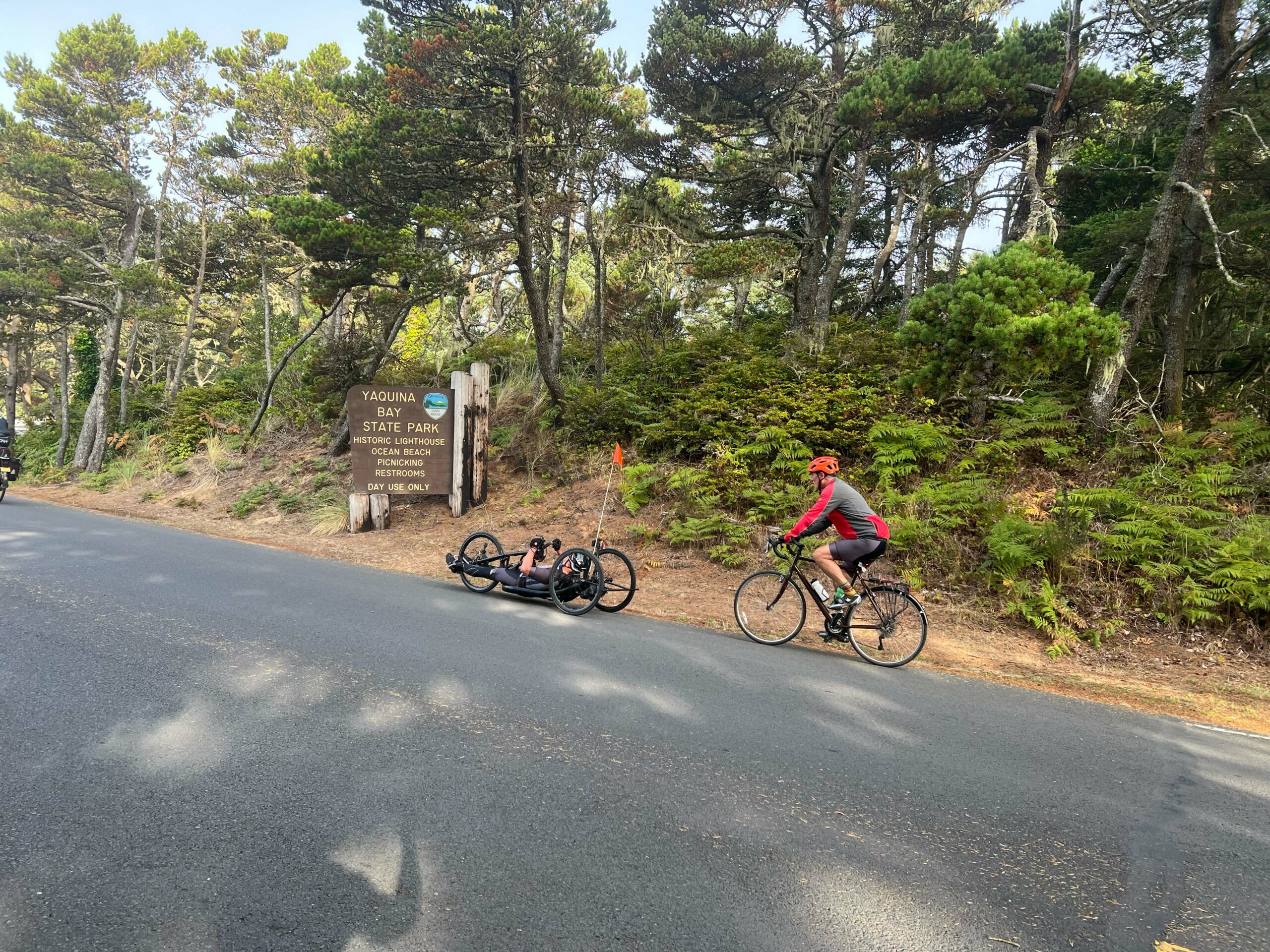 Biking uphill, an adaptive athlete in a hand-pedal bike leading with an OAS volunteer behind. Trees and shrubs in the background with a state park wooden sign shown.