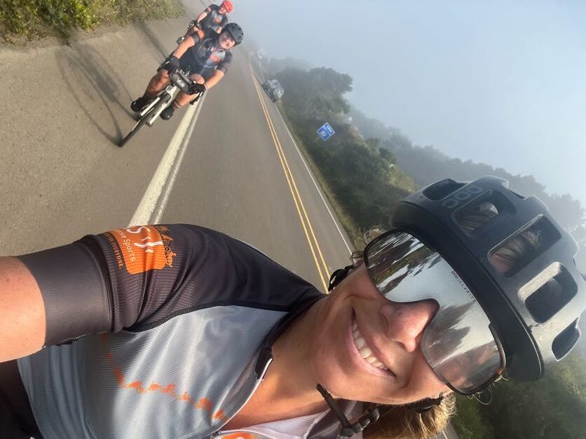 Selfie of as OAS staff leading the biking along the road with other OAS team members behind. There is fog visible in the skyline