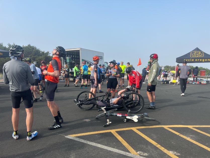 Large community group shot including other Cycle Oregon participants at a meeting location along the route in a parking lot. Approximately 20+ people shown in the photo with a Cycle Oregon booth tent visible in the far right corner.