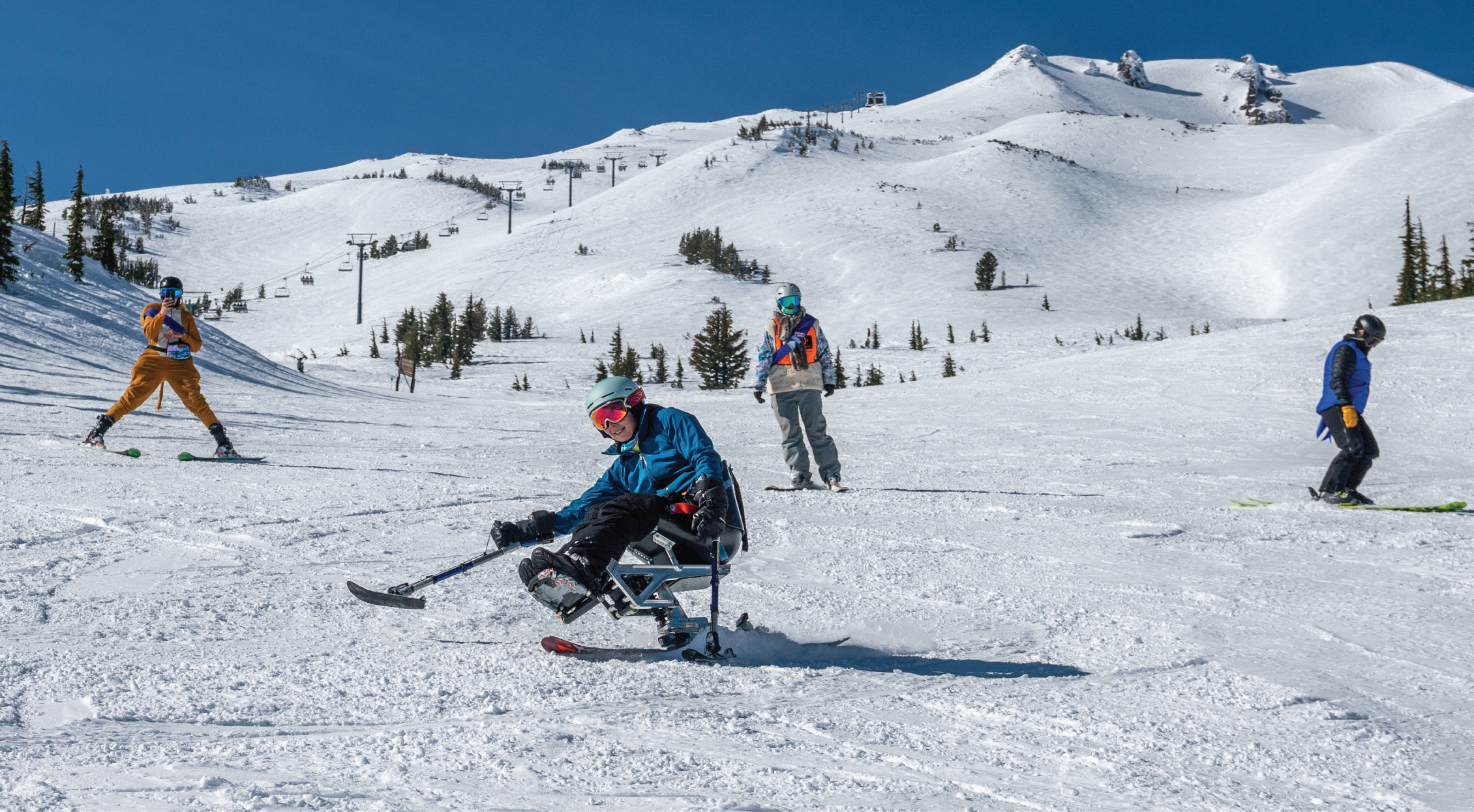 sit skier upfront gliding down the slope with instructor and two volunteers in the background. Mountains summit is visible in the far back