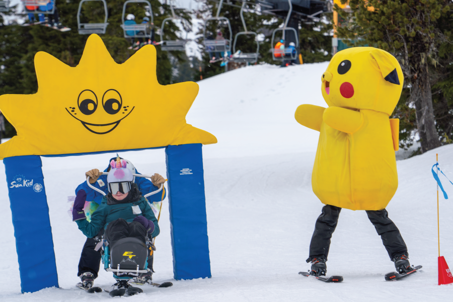 three people in the image, two are skiing under a foam course obstacle, one is an OAS instructor and another a young athlete in a bi-ski. Beside them cheering is someone in a giant Pikachu mascot costume, which is a yellow mouse-like cartoon character.