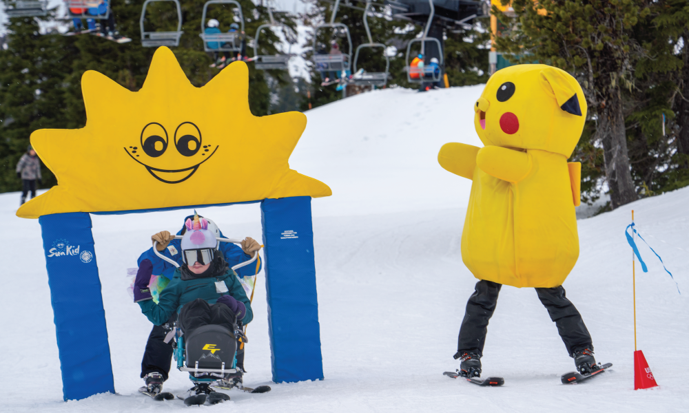 three people in the image, two are skiing under a foam course obstacle, one is an OAS instructor and another a young athlete in a bi-ski. Beside them cheering is someone in a giant Pikachu mascot costume, which is a yellow mouse-like cartoon character.