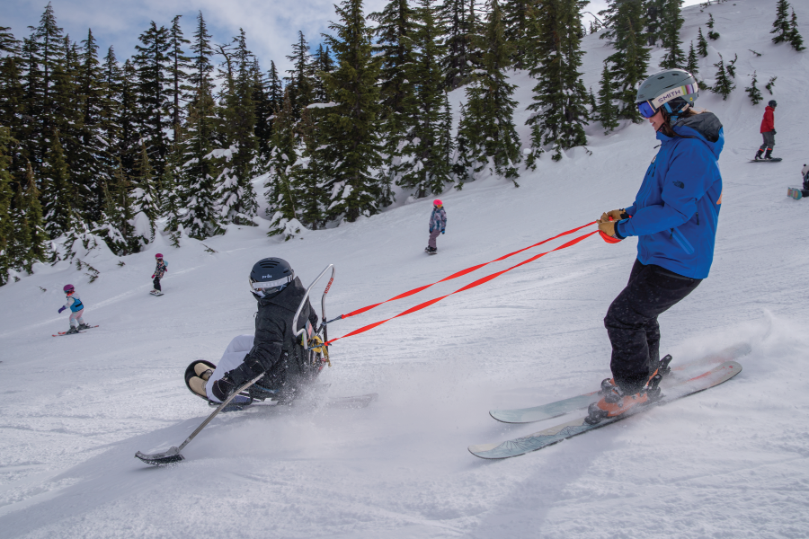 OAS Instructor with an adaptive athlete in a sit-ski, bi-ski as they descend down the mountain. In the background the treeline is visible with powder coming up behind their skis.