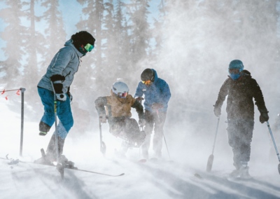 snow swirling around adaptive athletes at the top of the hill. there's two 3-track skiers and one mono-skier.