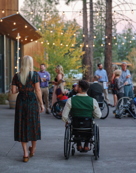 outside of the venue in the background the stationary adaptive bike set-up is going. in the foreground of the image are two attendees, one walking in a dark green dress with mid-length blonde hair and beside her is an adaptive athlete in a wheelchair in a white button-down and green satin vest.