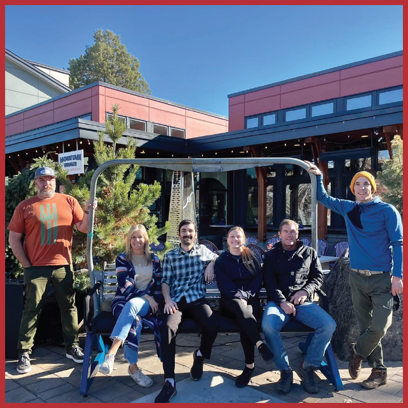 A group of 6 people, 4 sitting and 2 standing, beside a chairlift that is now a bench in front of Mountain Burger in Bend, OR. Sunny skies and green bushes in the background.