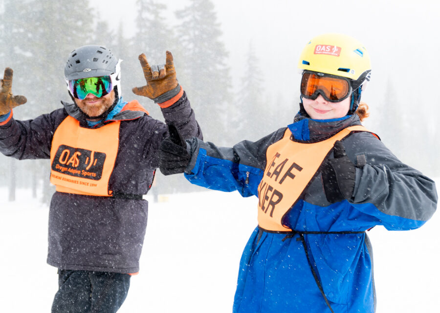 Outside on the snow, CamPAH coach in his helmet and googles holding up the 'I love you' icon sign to the camera. Next to him is a CamPAH skier in googles smiling with a thumbs up.