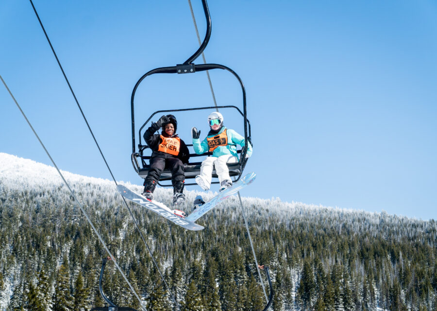 A CamPAH coach in their bright orange OAS bib with their camper wearing a Deaf Skier bib. Both are snowboarders waving from above on the chairlift.