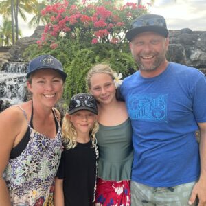 Josh with his family, two daughters and wife, behind them a tropical setting of a small waterfall and flora