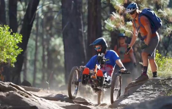 Jack in an adaptive mountain bike barreling down a trail with a full-cover helmet and OAS staff to his side ready to assist.