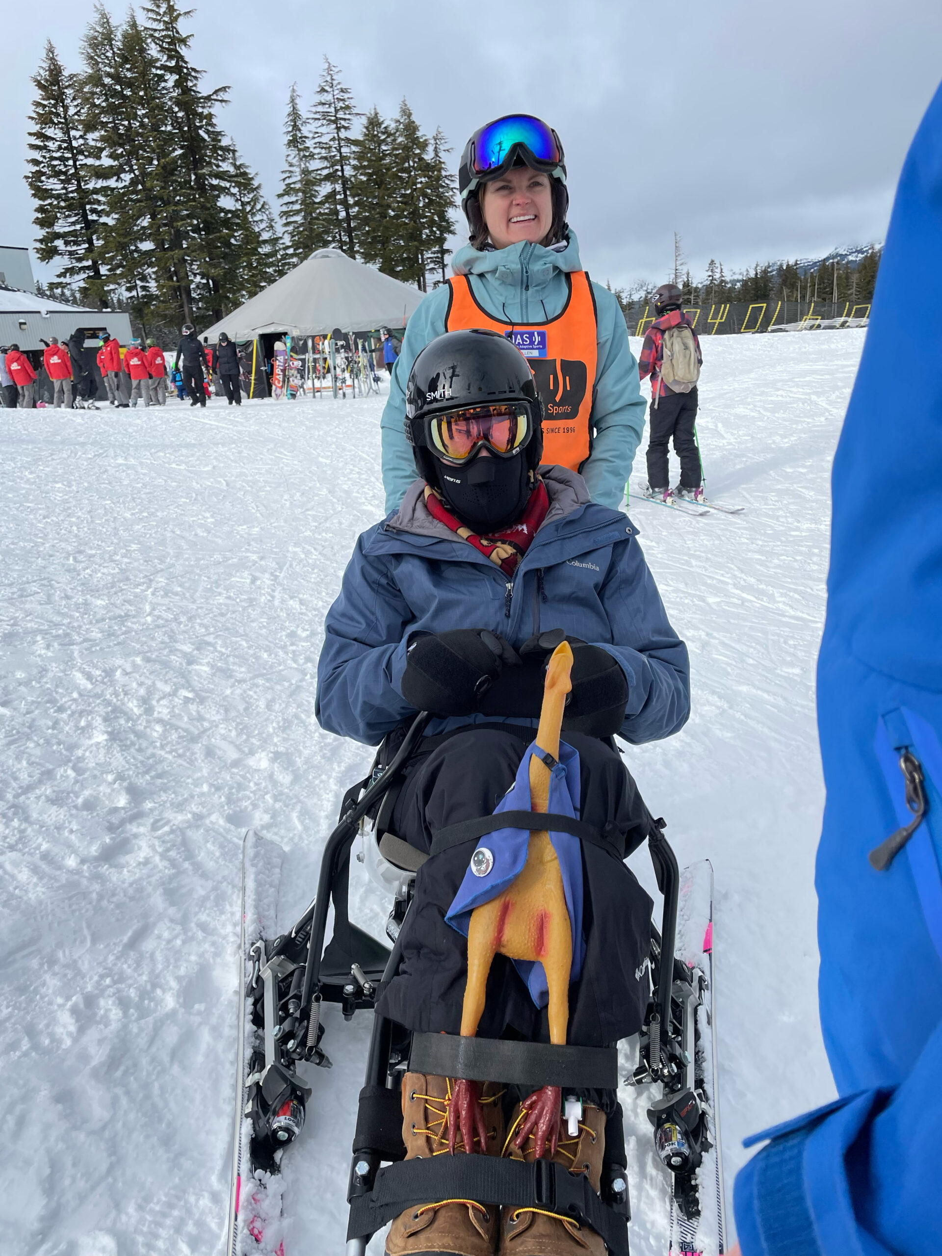 Jack in a sit-ski, an OAS volunteer standing behind him. His rubber chicken is attached to the front of his shins on the ski.