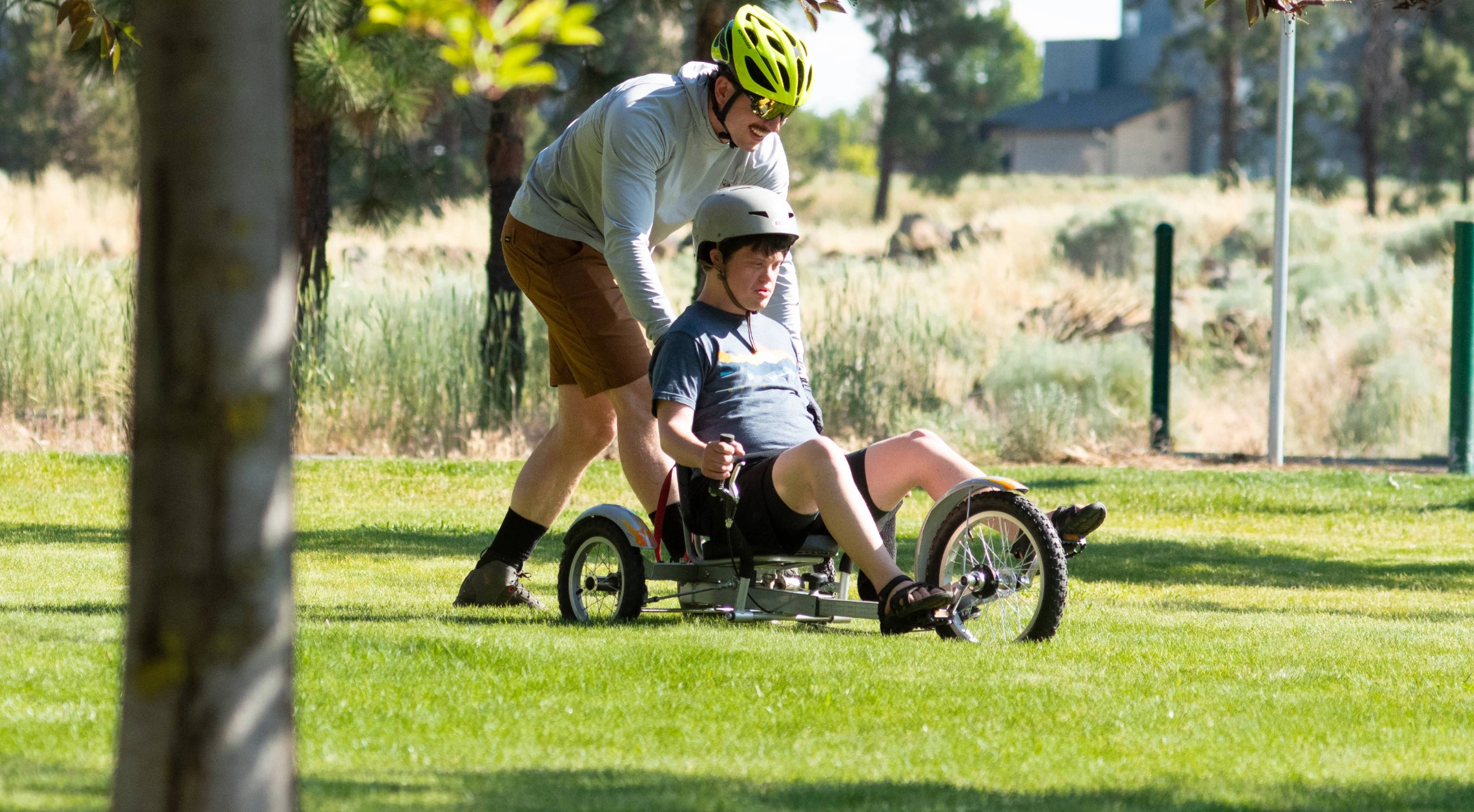 OAS volunteer in a green helmet supports a young adaptive athlete in a recumbent bike. bright green grass and greenery around them.