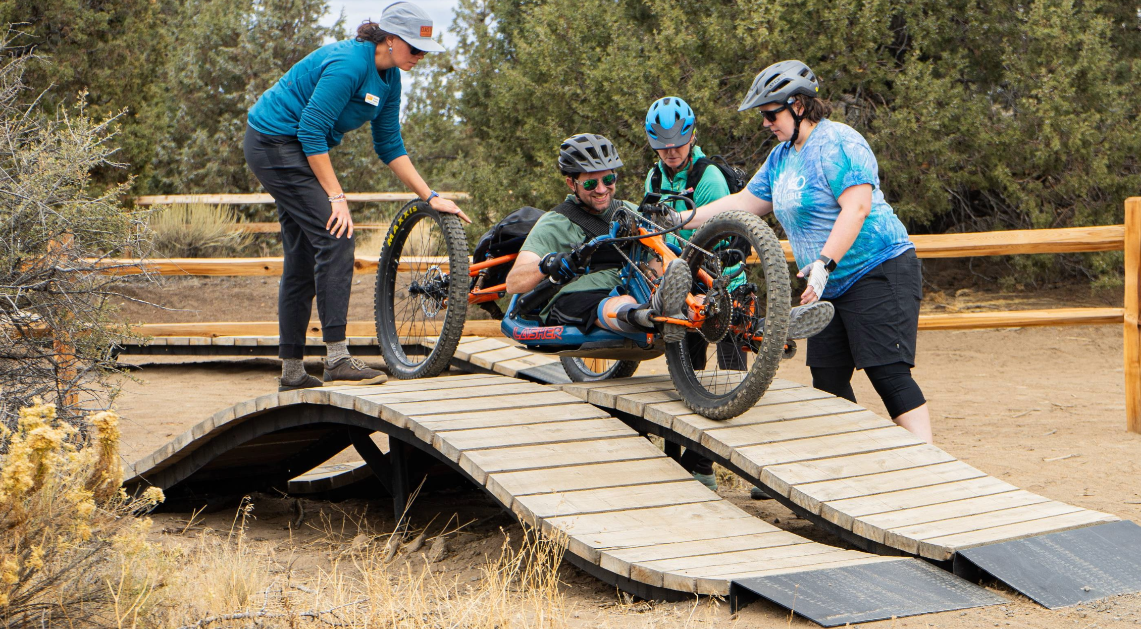 Three OAS volunteers support an adaptive athlete in an aMTB hand-cycle on a wooden ramp as they navigate balance. The athlete's smile visible.