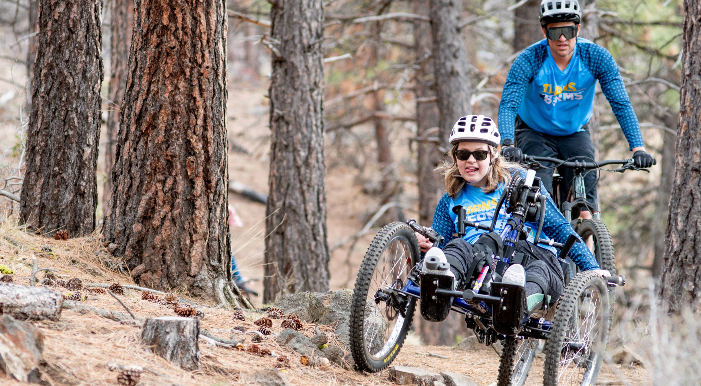 Female adaptive athlete in an aMTB hand-cycle powers through some technical terrien with an OAS volunteer on a standing bike following behind