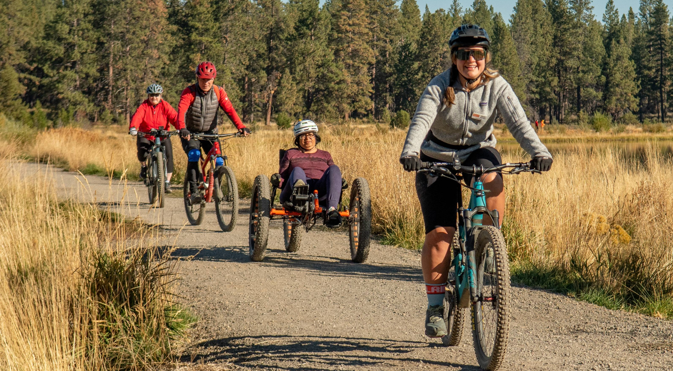 Three OAS volunteers guide an adaptive athlete in an off-road recumbent bike down a gravel path lined with tall dry grass