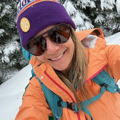 Sara in a purple beanie, sunglasses, and a light orange jacket out on a snowy sunny day.