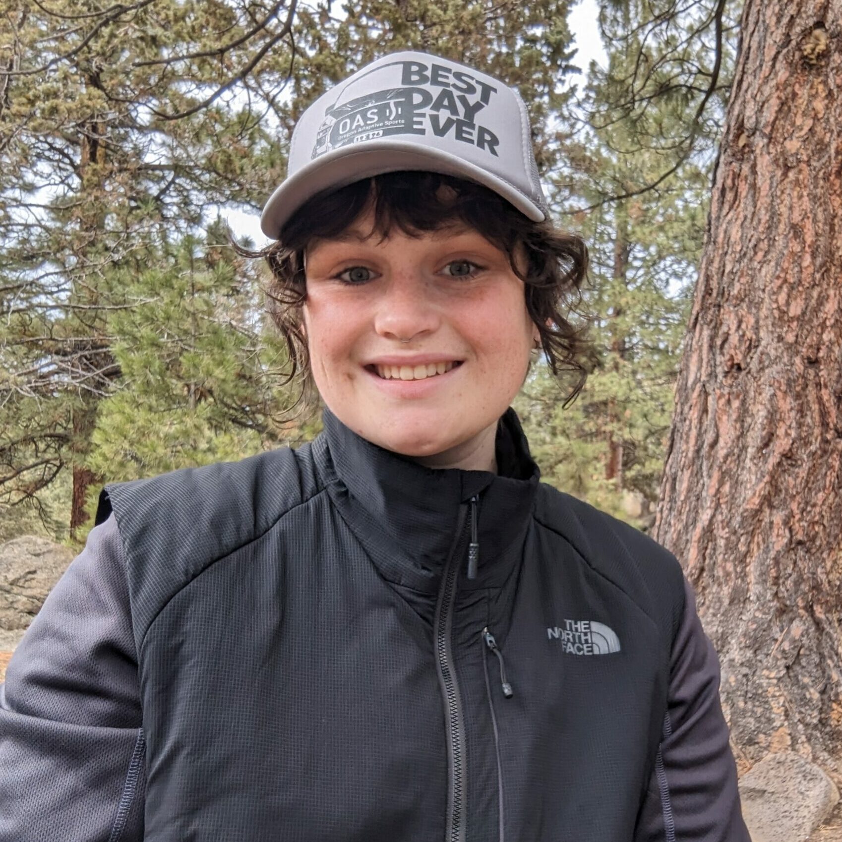 Samy wearing a grey hat, brown hair in a ponytail with pine trees in the background.