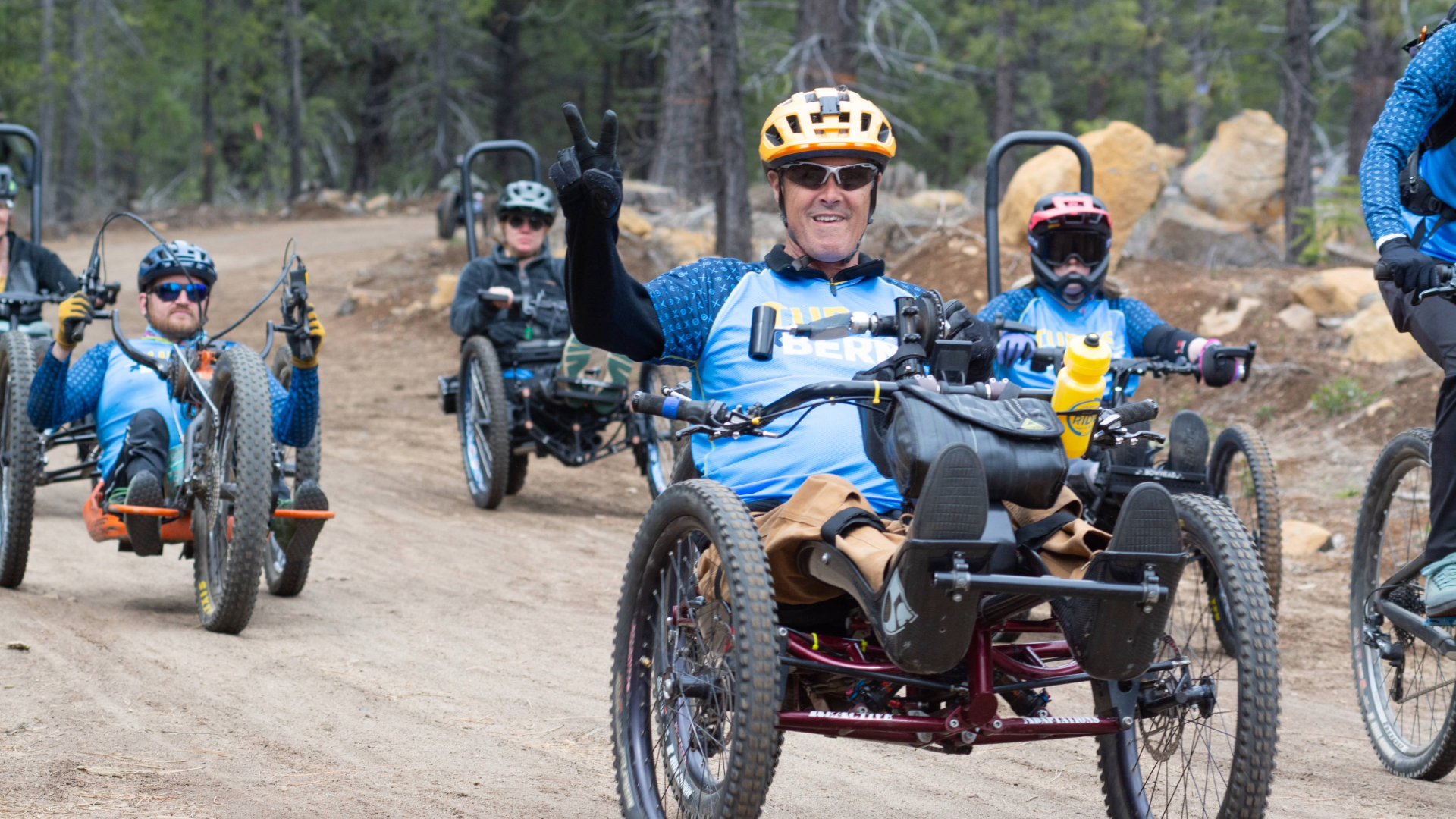 several athletes in aMTB handcycles going down the start of a trail, one athlete holds up a peace sign for the camera.