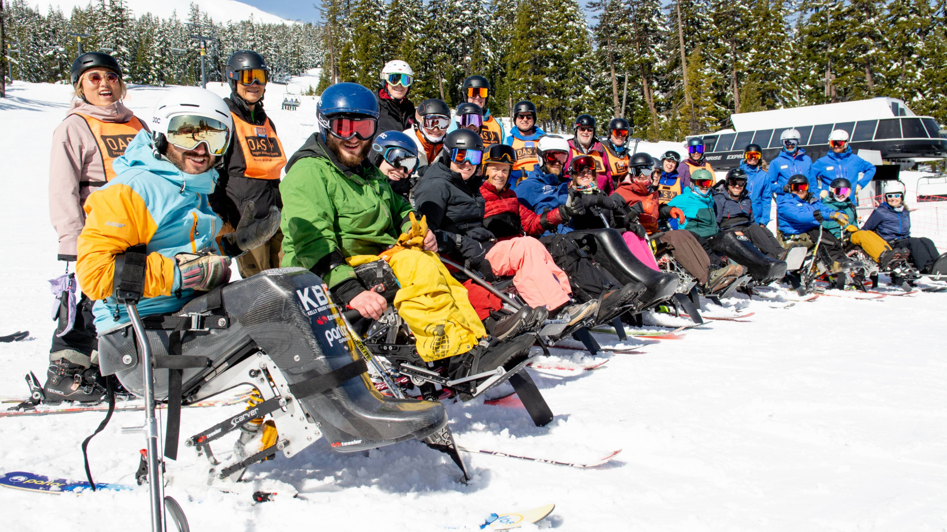 a group of 20 sit-skiers pose for a photo in a line with OAS volunteers and staffs stand behind them all smiling on a bright sunny day.