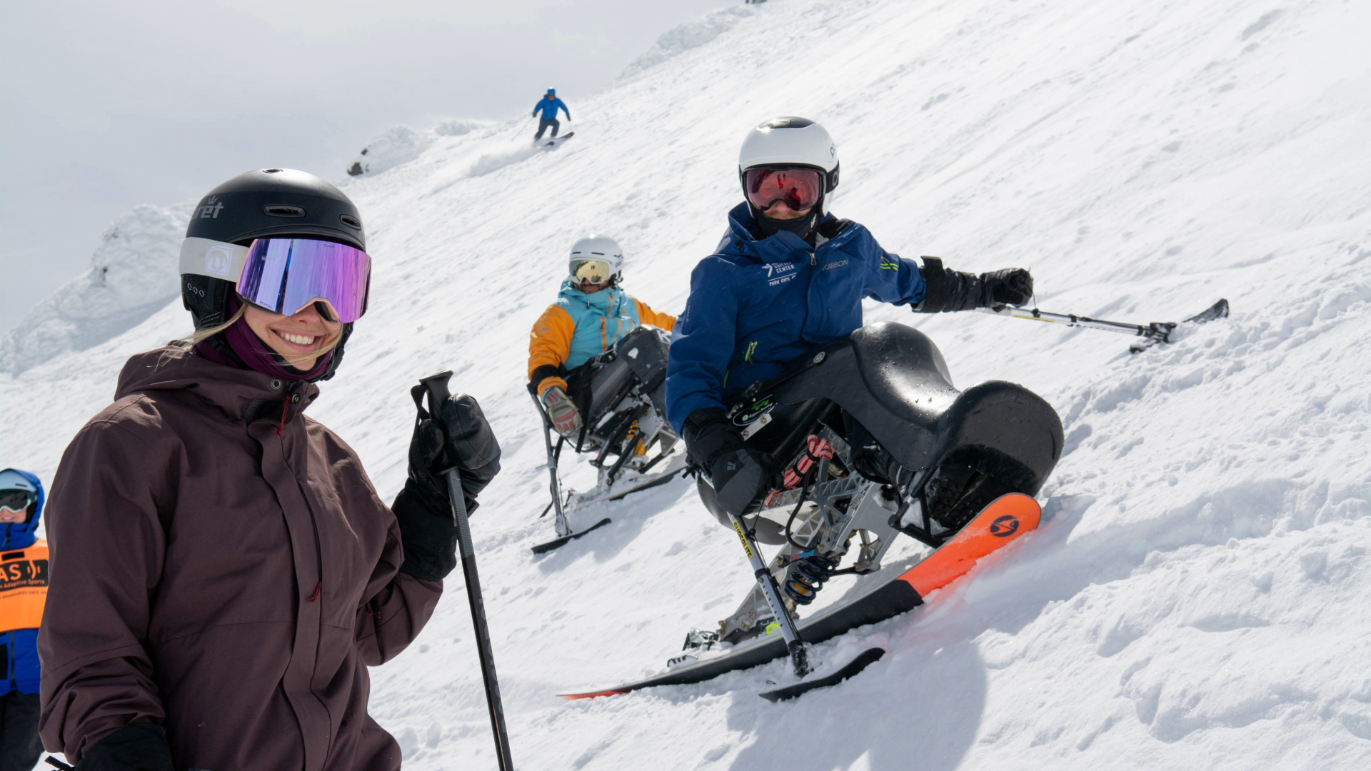 A 2-track skier supporting their partner in a sit-ski smiles at the camera with close behind them their partner and another sit-skier are stopped and angled on the steep slope. 