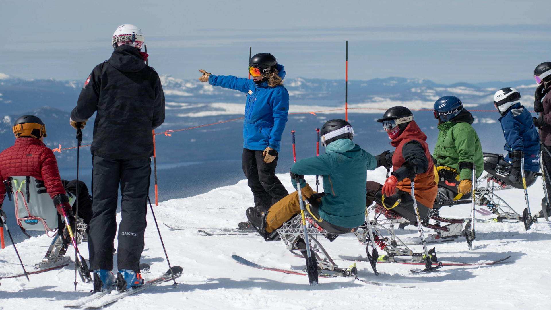 a handful of sit-skiers look to OAS Staff member on skis as they give direction at the top of summit. mountains visible in the distance from the panoramic view