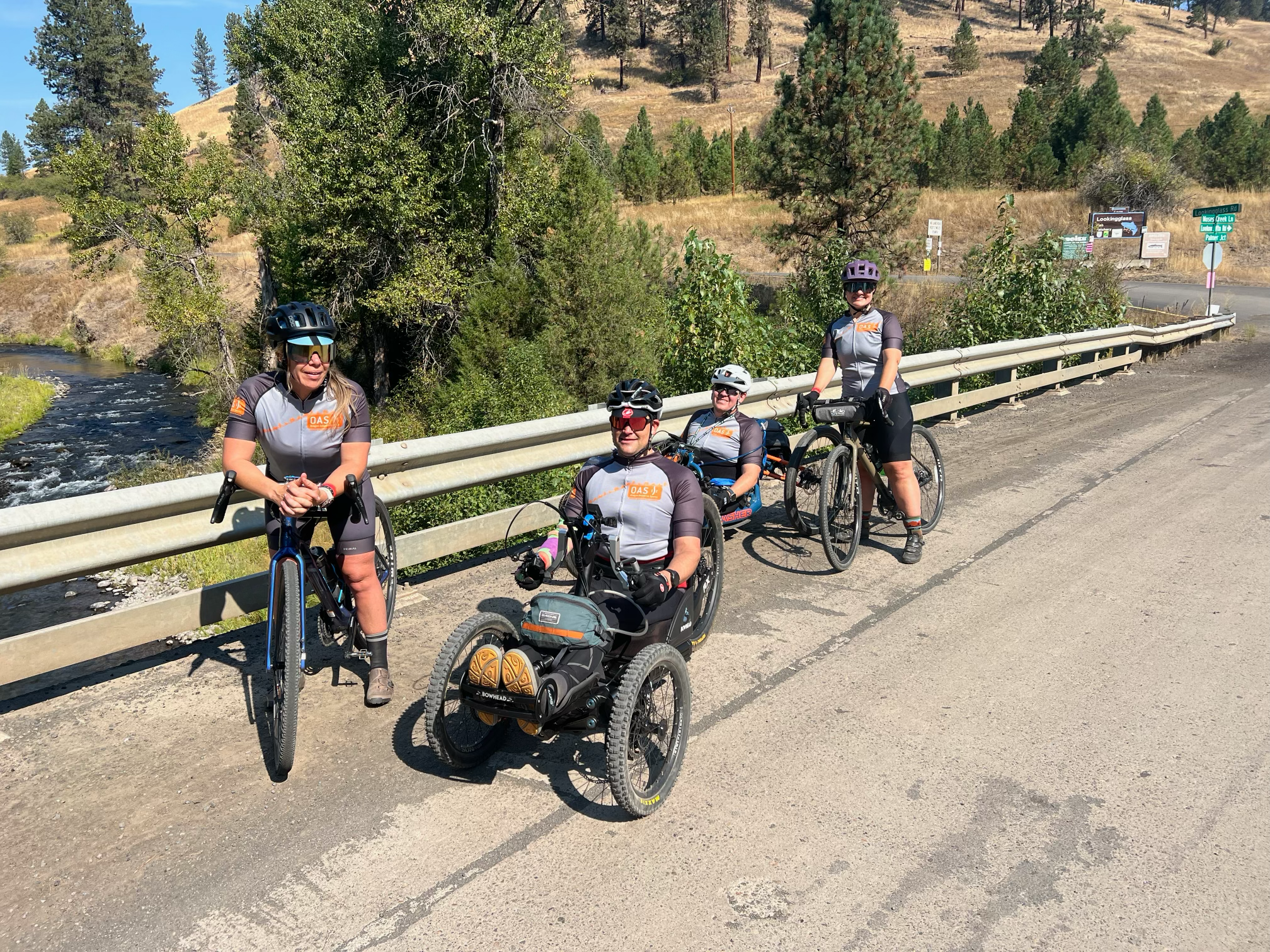 posed beside a metal rail, the road going over a small river, two OAS staff members on stand-up bikes with two athletes in handcycles smile at the camera.