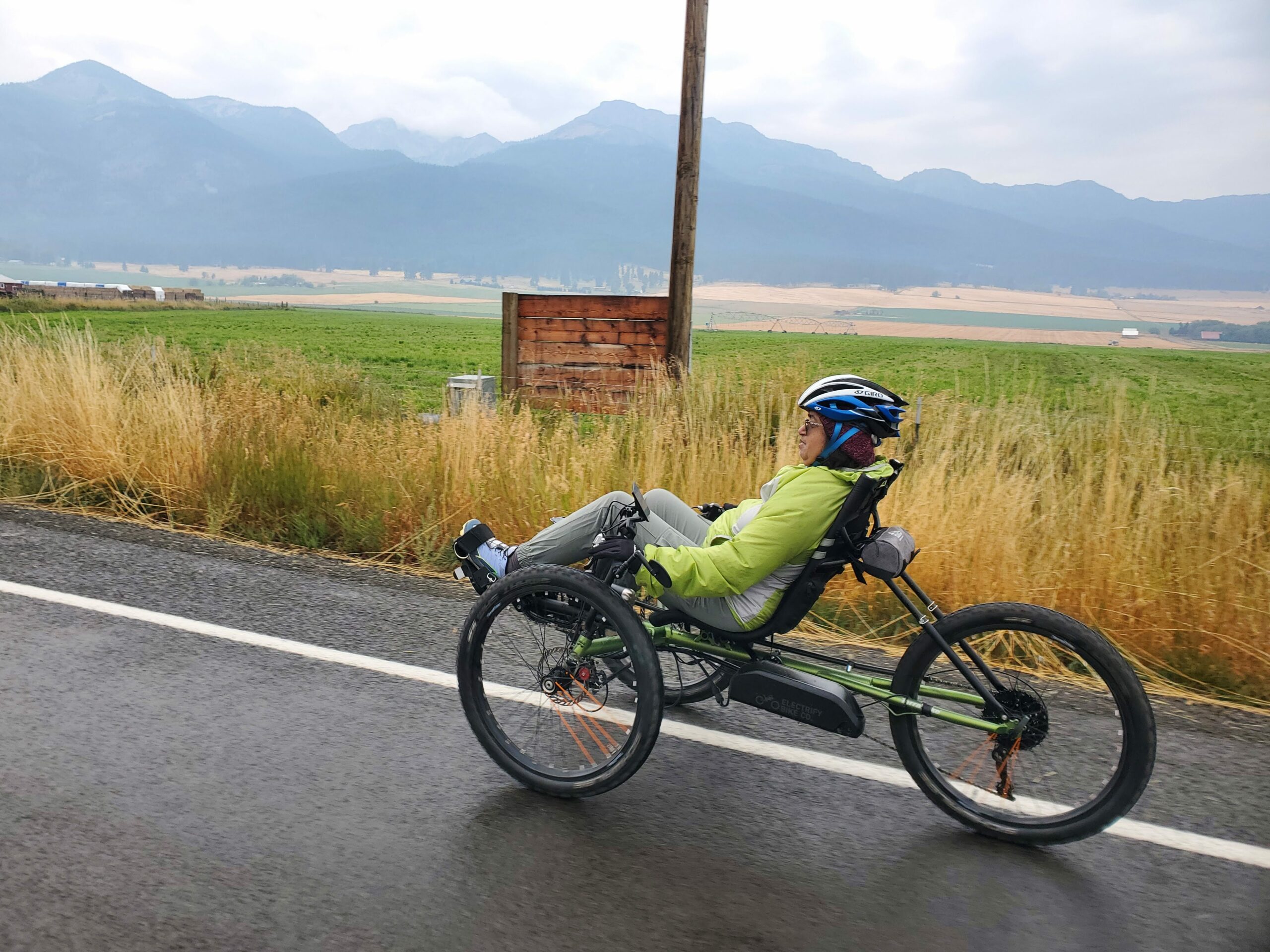 female in a recumbent cycle, mountains in the far distance with farmland behind the road. cloudy