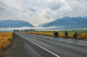 a wide angle landscape photo showing the mountains and farmland along the road. In the distance a pair on a tandem cycle is visible