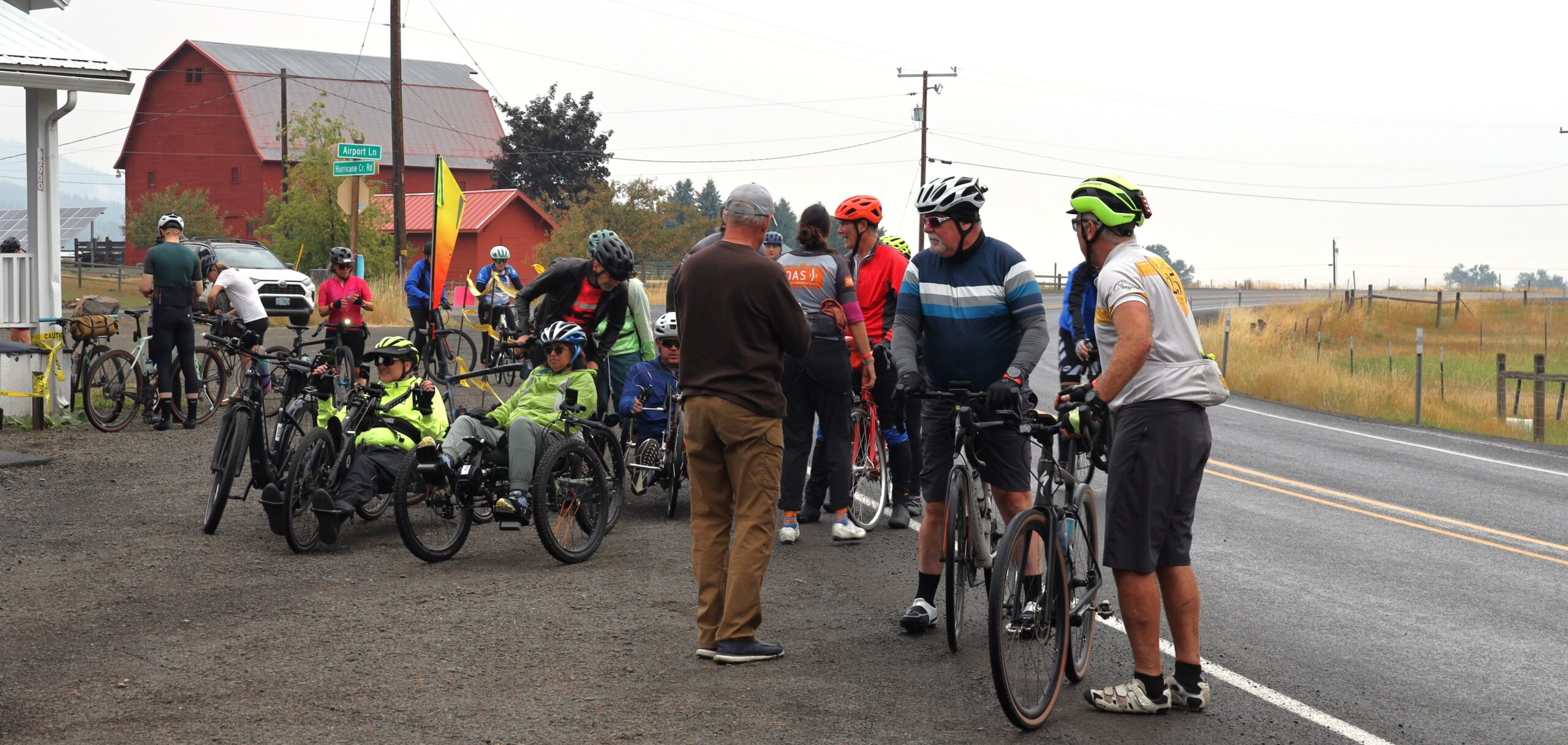 A pit stop during a cycling day. There's several people standing and some OAS athletes in recumbent and handcycles behind. The road is beside them from pulling off and a red barn visible in the distance