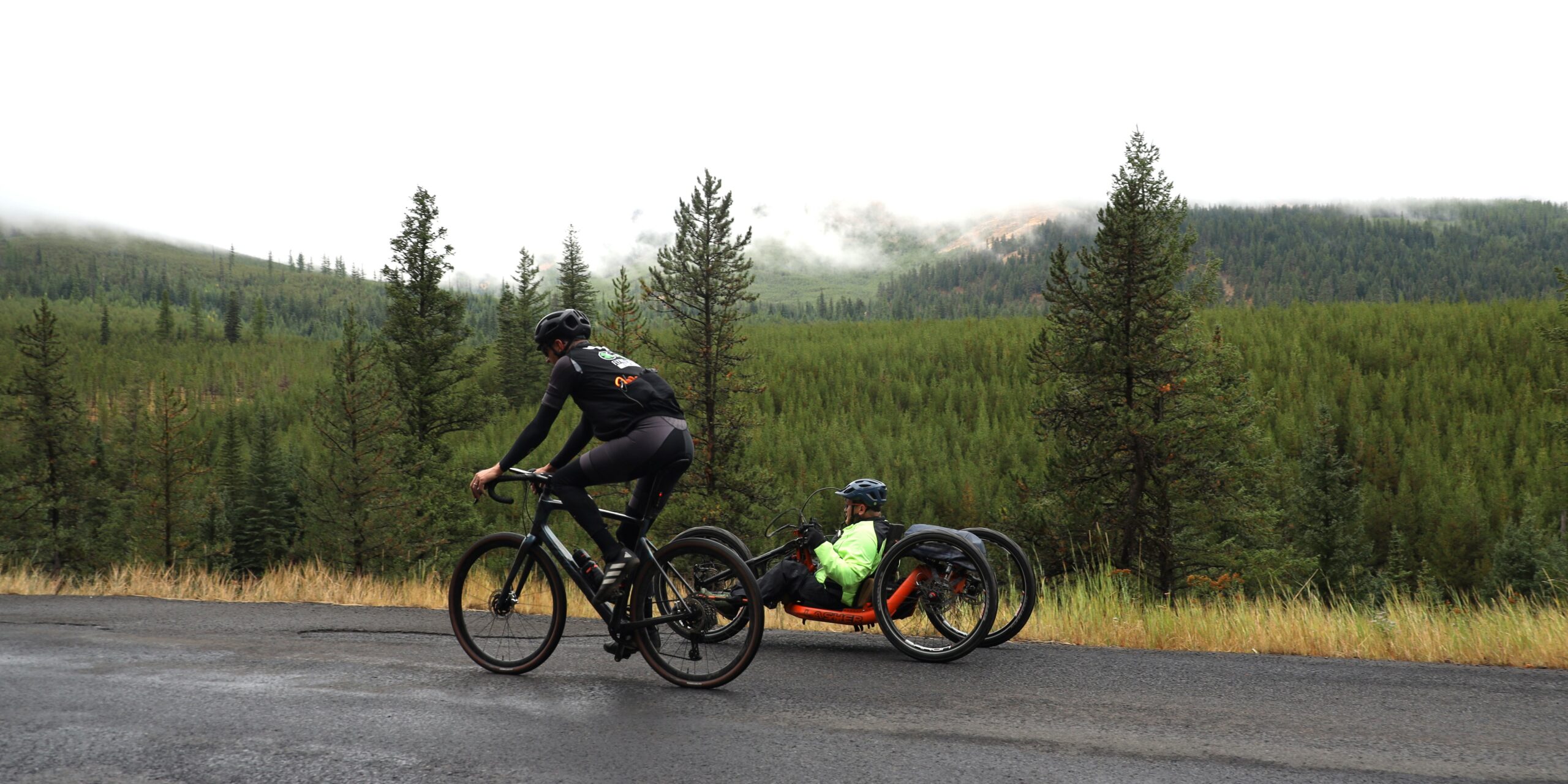 An OAS volunteer bikes beside an athlete in a handcycle, cloudy skies create an overcast on the photo with pine trees lining the background