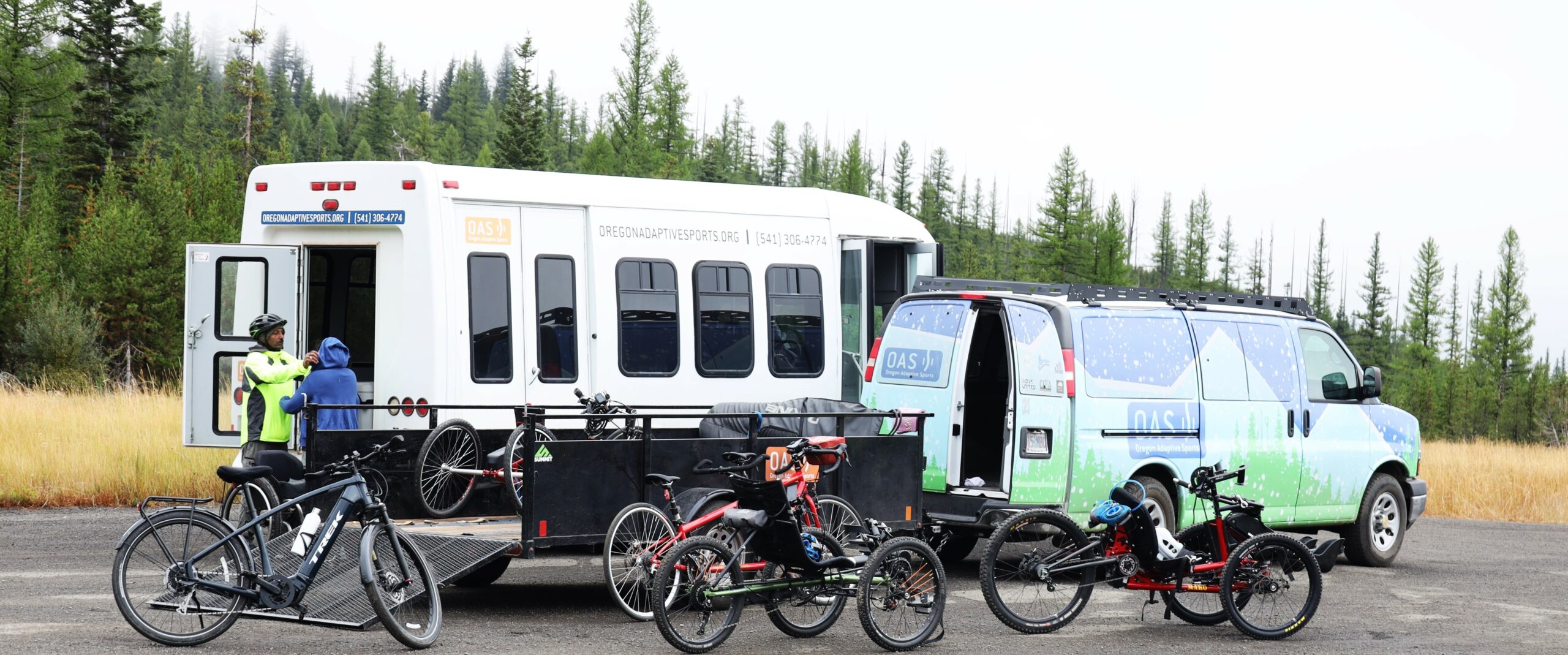 The 12-passenger OAS white bus with a pair from the OAS crew at the end of it, with the OAS van hauling a trailer positioned in front of the bus. Bikes are staged around