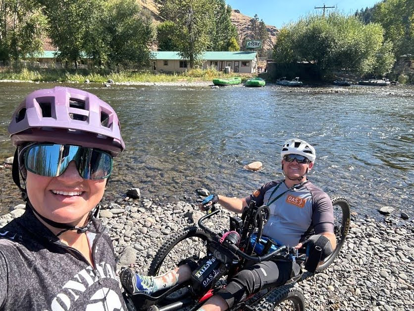 OAS staff smiles at the camera from the bottom frame with an athlete in a handcycle behind. both taking a break on a rocky shore beside a river