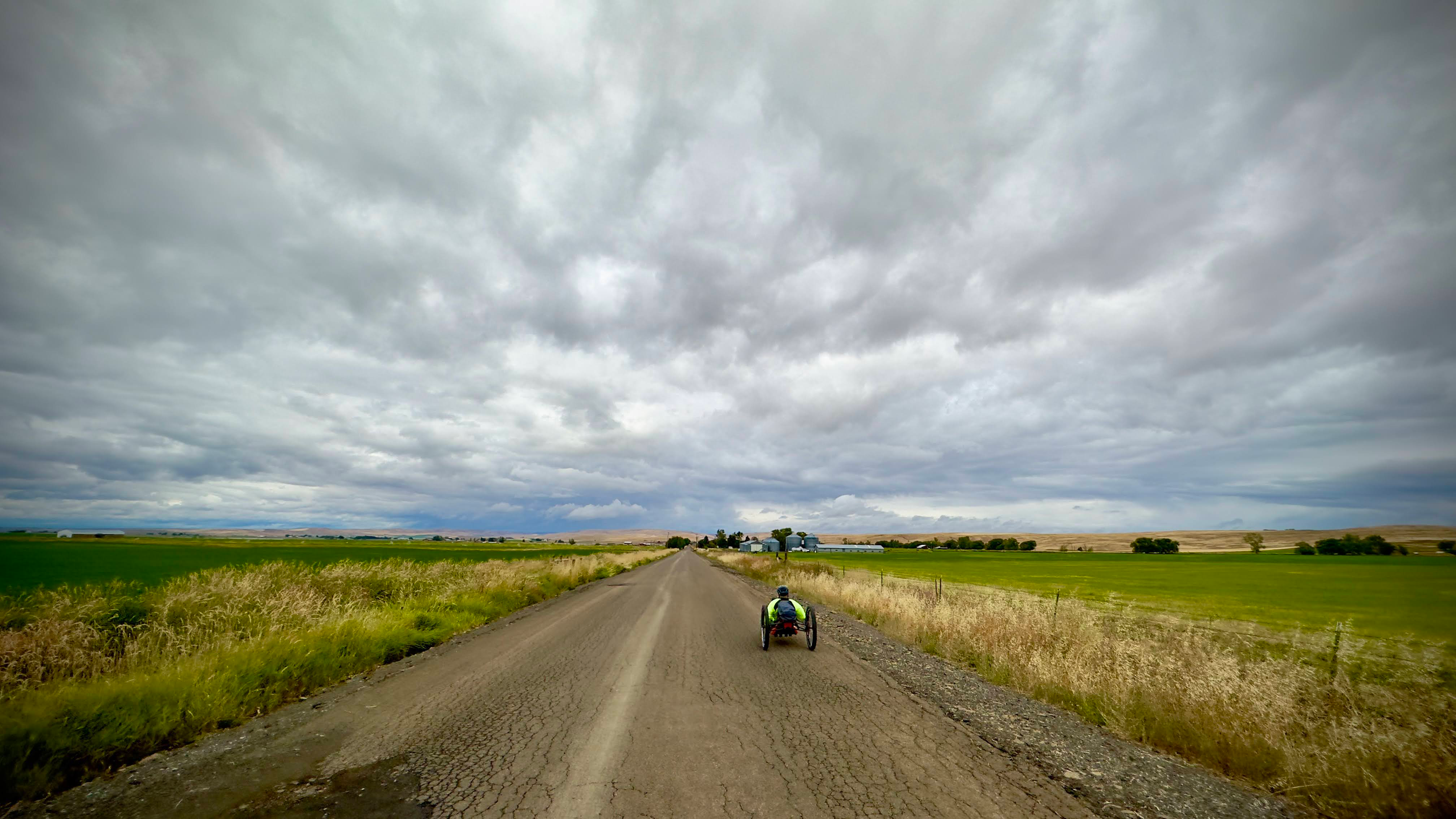 gray clouds cover the sky with the vast, farmland plains lining the old road with an athlete in a handcycle featured solo in the picture.