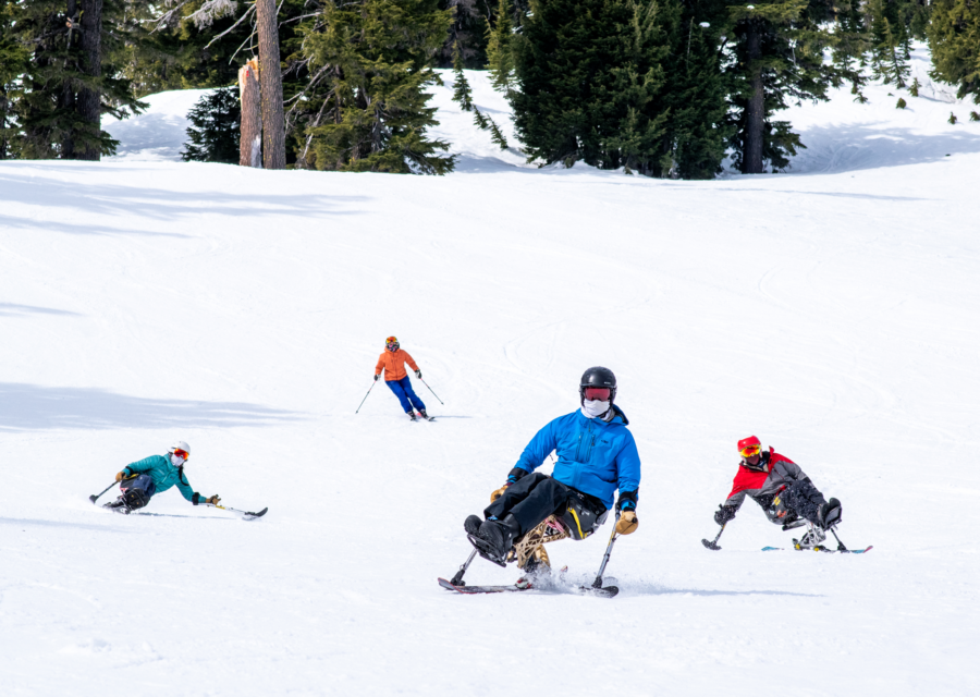 Three sit-skiers turning down the slope with a stand-up skier behind.